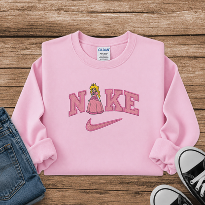 A pink long-sleeve sweatshirt laid flat on a rustic wood background, featuring a 'Nike' wordmark with a princess peach character in a pink dress and a pink swoosh. A Gildan brand tag is visible inside the collar.