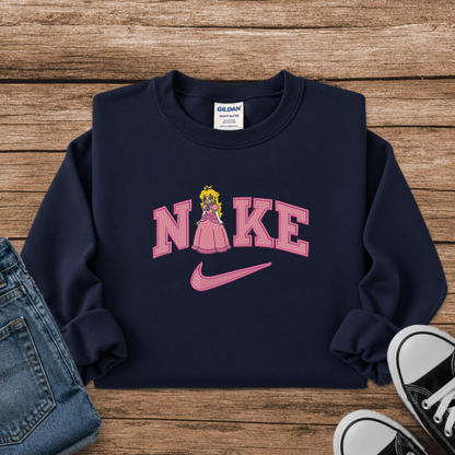 A navy blue crewneck sweatshirt with a pink 'NIKE' graphic featuring a princess character and a pink Nike swoosh, styled in a flat lay with jeans and sneakers on a wooden background.