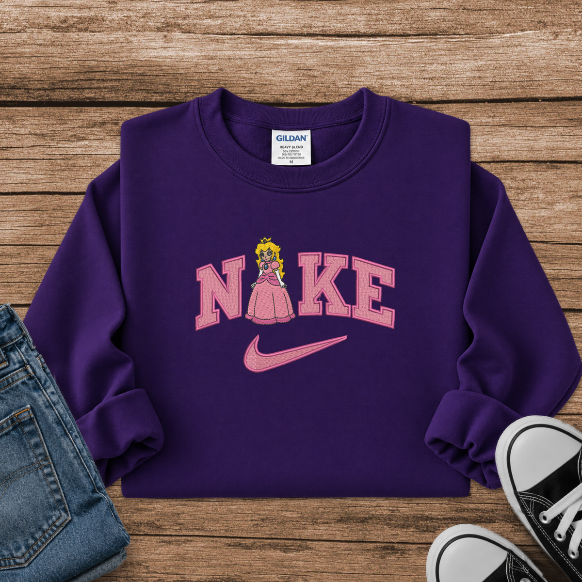 A purple crewneck sweatshirt styled in a flat lay on a wooden surface, featuring a pink 'NIKE' graphic with a princess character and a pink swoosh. Blue jeans and black sneakers are partially visible as styling props.