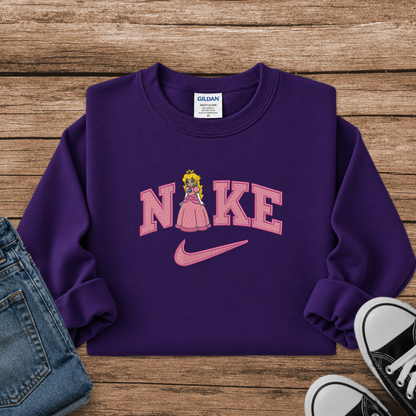 A purple crewneck sweatshirt styled in a flat lay on a wooden surface, featuring a pink 'NIKE' graphic with a princess character and a pink swoosh. Blue jeans and black sneakers are partially visible as styling props.
