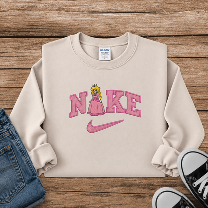 A beige crewneck sweatshirt styled in a flat lay on a wooden surface, featuring a pink 'Nike' graphic with a princess character and a pink swoosh. Blue jeans and black sneakers are partially visible as styling props.