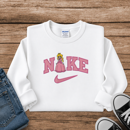 A white long-sleeve sweatshirt laid flat on a rustic wood background, styled with blue jeans and black sneakers. The sweatshirt features a pink graphic spelling 'NIKE' with a pink Nike swoosh and a small princess peach character replacing the 'I'.