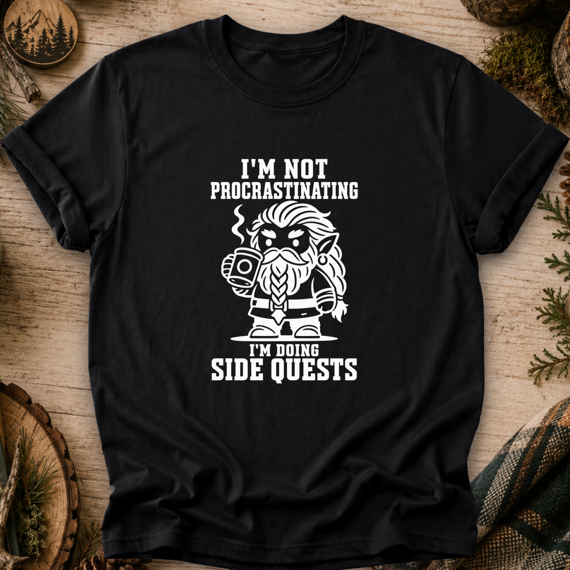 A black short-sleeve graphic T-shirt laid flat on a rustic wood surface with pine branches and decorative props around it. The shirt has a large white printed graphic and text reading, "I'M NOT PROCASTINATING I'M DOING SIDE QUESTS" with a stylized bearded character holding a mug.