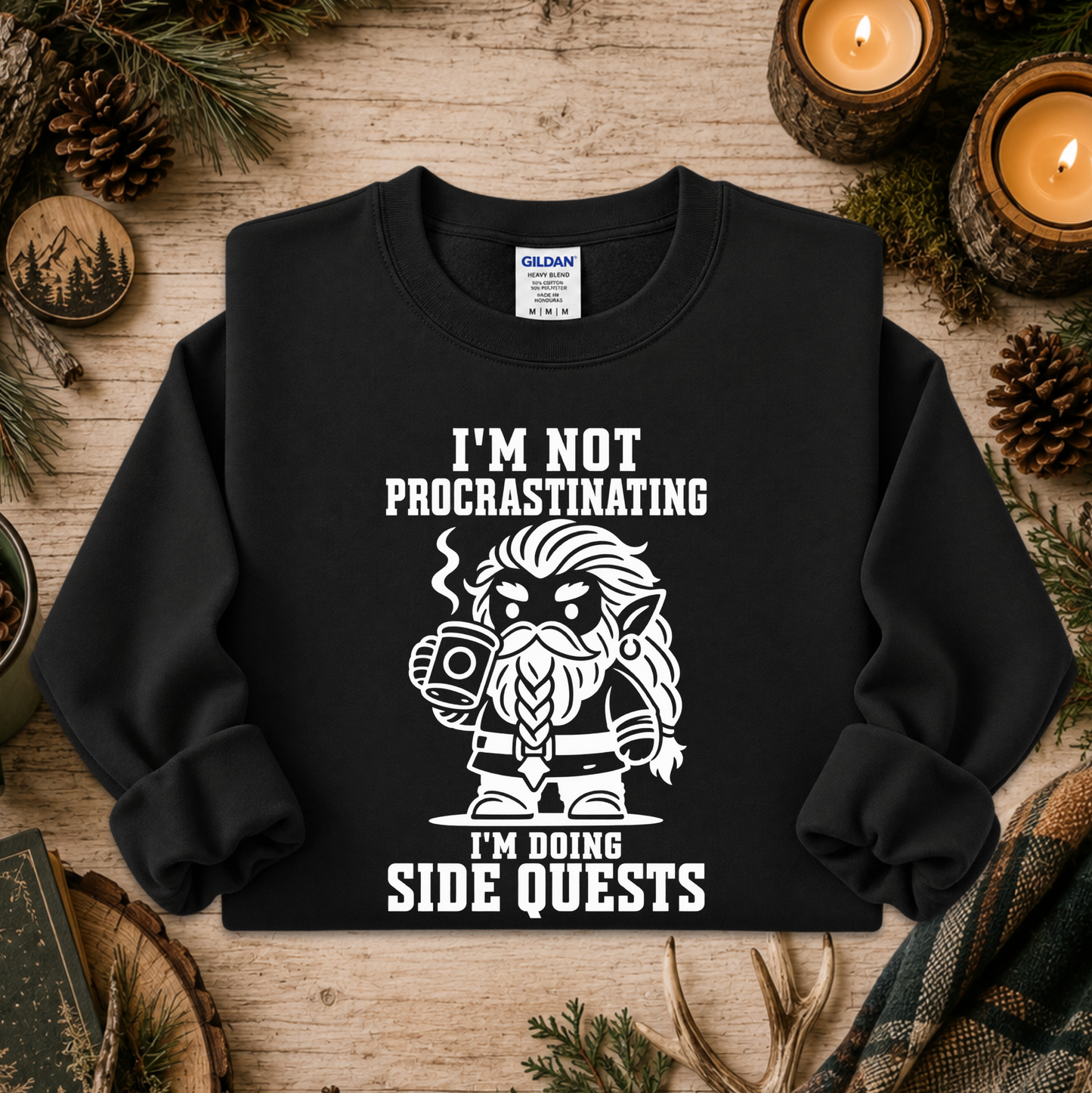 A black crewneck sweatshirt with a white graphic of a bearded elf character holding a mug, surrounded by the text 'I'M NOT PROCASTINATING I'M DOING SIDE QUESTS'. The sweatshirt is displayed in a flat-lay setting on a rustic wooden surface with pine needles, pinecones, and lit candles.