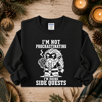 A black crewneck sweatshirt with a white graphic of a bearded elf character holding a mug, surrounded by the text 'I'M NOT PROCASTINATING I'M DOING SIDE QUESTS'. The sweatshirt is displayed in a flat-lay setting on a rustic wooden surface with pine needles, pinecones, and lit candles.