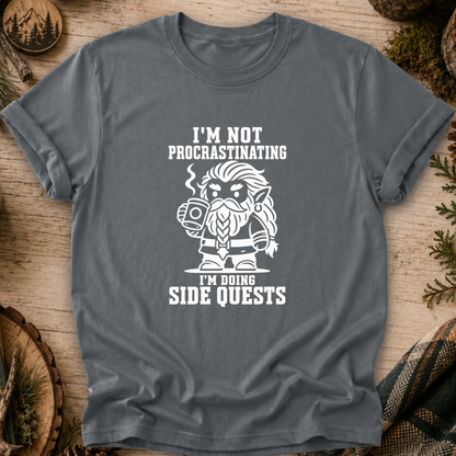 A dark gray graphic t-shirt laid flat on a rustic wooden background with pine needles and wood slices. The shirt features a white graphic of a bearded fantasy character holding a mug, with the text 'I'M NOT PROCRASTINATING' above and 'I'M DOING SIDE QUESTS' below.
