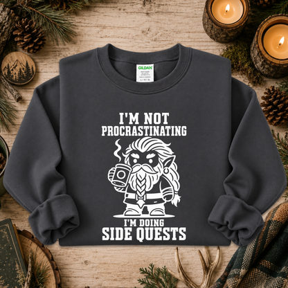 A dark charcoal gray crewneck sweatshirt with a white graphic of a bearded fantasy character holding a mug, surrounded by the text 'I'M NOT PROCRASTINATING I'M DOING SIDE QUESTS'. The sweatshirt is laid flat on a rustic wooden surface decorated with pine needles, pinecones, and lit candles.