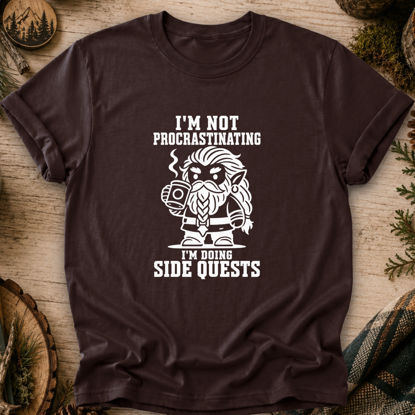 A black short-sleeve graphic T-shirt laid flat on a rustic wood surface with pine branches and decorative props around it. The shirt has a large white printed graphic and text reading, "I'M NOT PROCASTINATING I'M DOING SIDE QUESTS" with a stylized bearded character holding a mug.