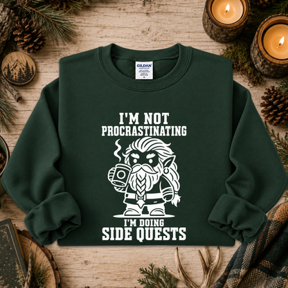 A dark green crewneck sweatshirt with a white graphic and text reading 'I'M NOT PROCASTINATING I'M DOING SIDE QUESTS' and a cartoon bearded character holding a mug, displayed on a rustic wooden surface with candles, pinecones, and evergreen branches.