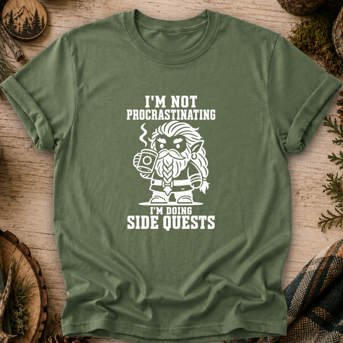 An olive green short-sleeve t-shirt with a white graphic print of a bearded fantasy character holding a mug, surrounded by the text 'I'M NOT PROCRASTINATING I'M DOING SIDE QUESTS'. The shirt is displayed in a flat-lay arrangement on a rustic wooden background with pine needles and wood slices.