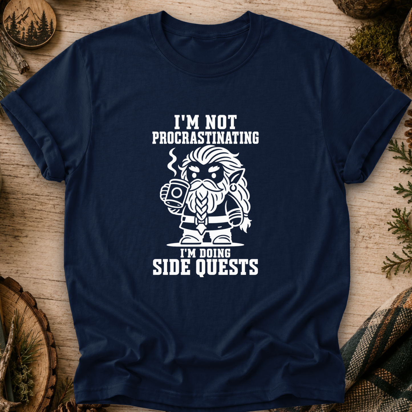 A navy blue short-sleeve t-shirt with a white graphic print of a bearded fantasy character holding a mug, surrounded by the text 'I'M NOT PROCRASTINATING I'M DOING SIDE QUESTS'. The shirt is displayed flat on a rustic wooden background with pine needles and wood slices.