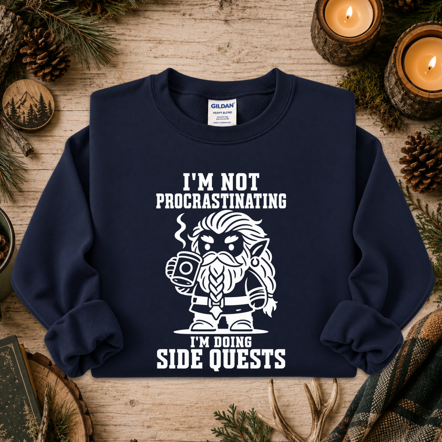 A navy blue crewneck sweatshirt with a white graphic of a bearded elf character holding a mug, surrounded by the text 'I'M NOT PROCRASTINATING I'M DOING SIDE QUESTS'. The sweatshirt is displayed in a flat-lay setting on a rustic wooden table decorated with pine branches, pinecones, and lit candles.