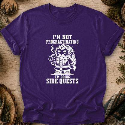 A purple short-sleeve t-shirt with a white graphic of a bearded dwarf character holding a mug, surrounded by the text 'I'M NOT PROCRASTINATING I'M DOING SIDE QUESTS'. The shirt is displayed flat on a rustic wooden background with pine needles and wood slices.