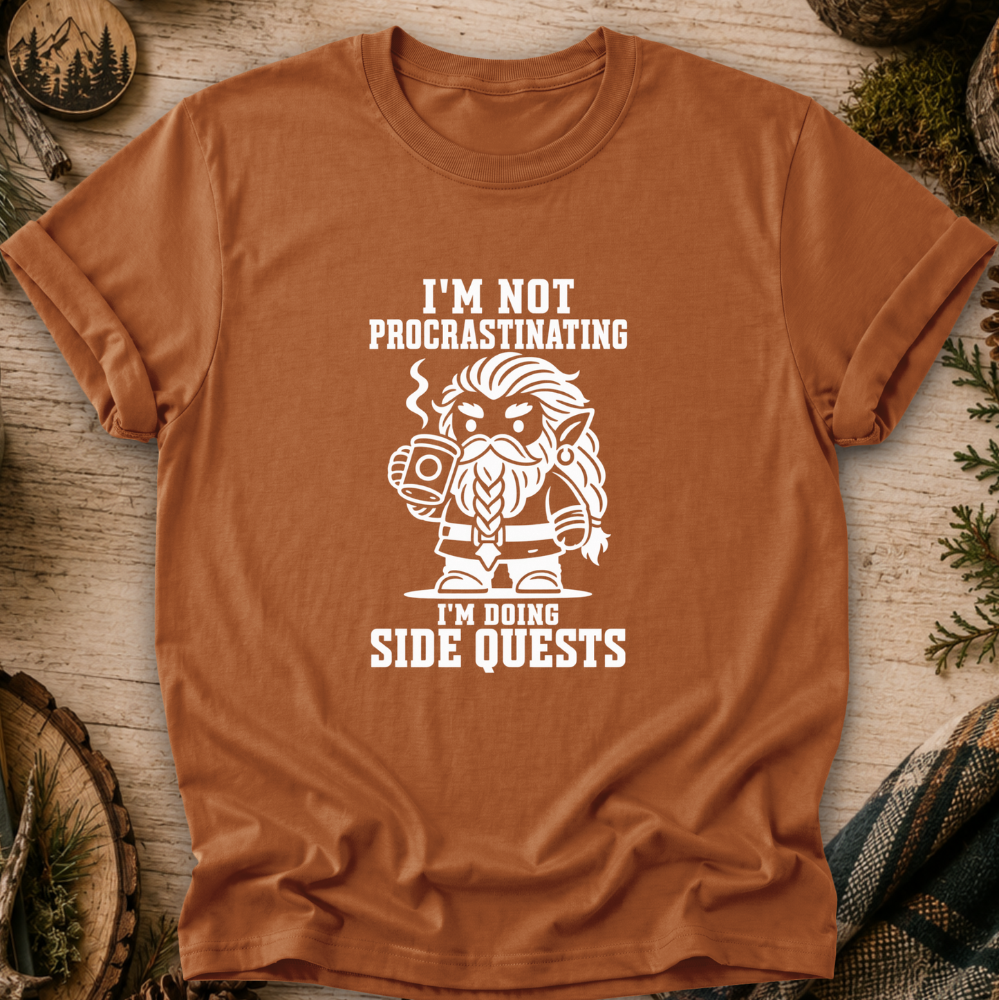 A rust-orange crewneck t-shirt with a white graphic of a bearded wizard holding a mug, surrounded by the text 'I'M NOT PROCRASTINATING I'M DOING SIDE QUESTS'. The shirt is displayed flat on a rustic wooden background with pine needles and wood slices.