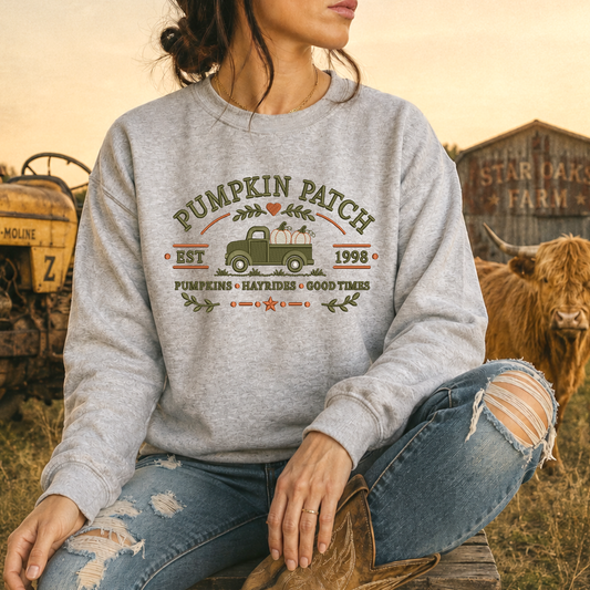 A woman wearing a heather gray crewneck sweatshirt with a 'Pumpkin Patch 1998' graphic, sitting in a rustic outdoor setting with a cow and farm equipment in the background.