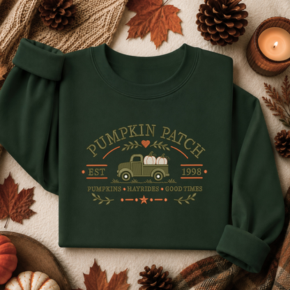 A dark forest green long-sleeve sweatshirt with a vintage-style autumn graphic reading 'Pumpkin Patch' and 'Est. 1998' around a truck carrying pumpkins, styled in a flat lay with fall leaves, pinecones, a candle, and a knit blanket.