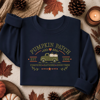 A navy blue long-sleeve sweatshirt laid flat on a neutral fabric background, surrounded by autumn props including pinecones, dried leaves, a candle, and a small pumpkin. The sweatshirt features a vintage-style graphic with the words “Pumpkin Patch,” a green pickup truck carrying pumpkins, “Est.