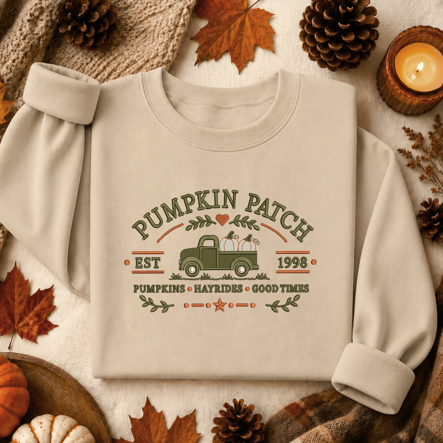 A cream-colored long-sleeve sweatshirt with a vintage-style 'Pumpkin Patch' graphic, laid flat on a neutral fabric background surrounded by autumn props like pinecones, leaves, and a candle.
