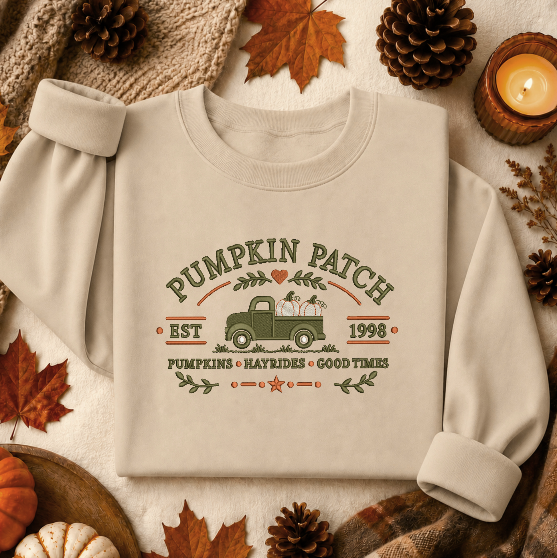 A cream-colored long-sleeve sweatshirt with a vintage-style 'Pumpkin Patch' graphic, laid flat on a neutral fabric background surrounded by autumn props like pinecones, leaves, and a candle.