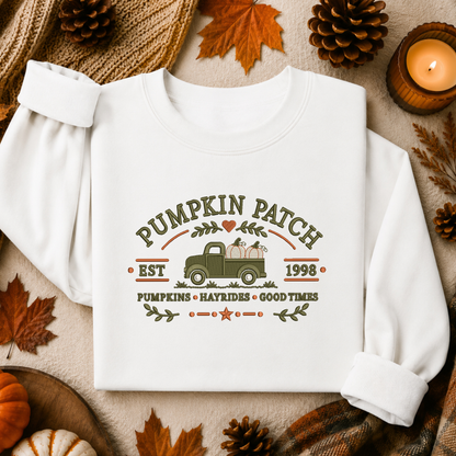 A white long-sleeve sweatshirt laid flat on a cozy autumn-themed background with pinecones, fall leaves, a candle, and small pumpkins. The sweatshirt has a vintage-style printed graphic reading “Pumpkin Patch” with a green pickup truck carrying pumpkins and the text “Pumpkins • Hayrides • Good Times” below.