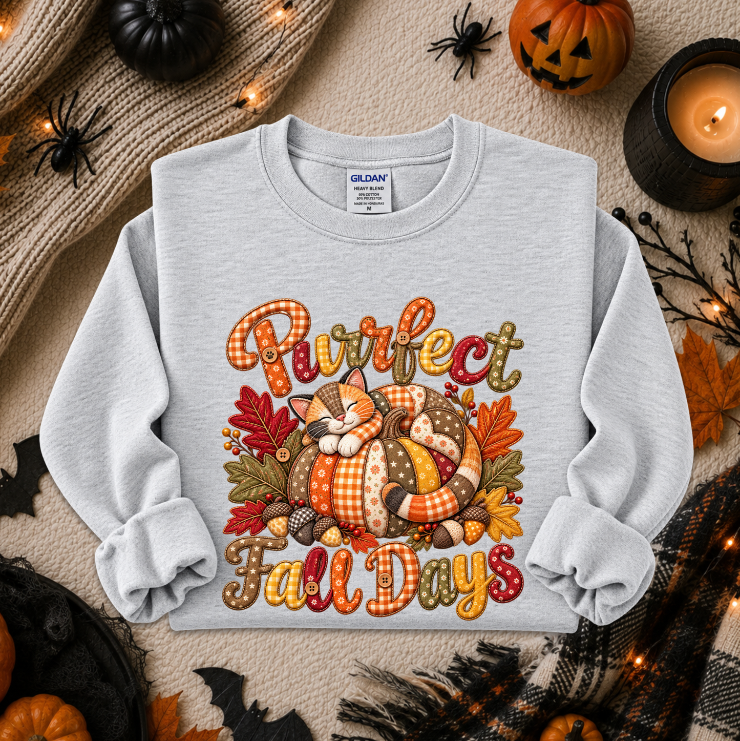 A light heather gray crewneck sweatshirt featuring a colorful graphic of a sleeping cat in a pumpkin with the text 'Purrfect Fall Days' in autumn-themed lettering. The sweatshirt is displayed in a flat-lay arrangement with seasonal props including a pumpkin, a candle, a scarf, and spider decorations on a textured beige background.