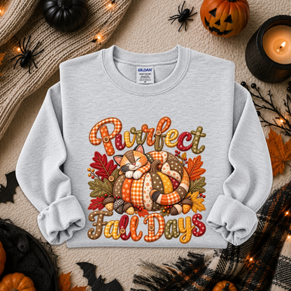 A light heather gray crewneck sweatshirt featuring a colorful graphic of a sleeping cat in a pumpkin with the text 'Purrfect Fall Days' in autumn-themed lettering. The sweatshirt is displayed in a flat-lay arrangement with seasonal props including a pumpkin, a candle, a scarf, and spider decorations on a textured beige background.