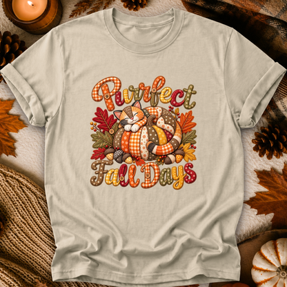 A light beige graphic t-shirt laid flat on a cozy autumn-themed background with pinecones, leaves, and a candle. The shirt features a colorful graphic of a sleeping cat curled up in a pumpkin with the text 'Purrfect Fall Days'.
