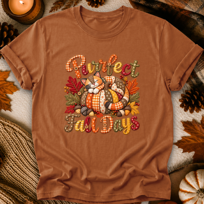 A brown short-sleeve t-shirt laid flat with an autumn-themed graphic that reads 'Purrfect Fall Days' around a sleeping cat and pumpkins. The shirt is styled with fall leaves, pinecones, a candle, and a knit blanket.