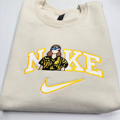A folded cream-colored sweatshirt featuring a custom graphic of the word 'NIkE' in yellow and white varsity letters, with a stranger things eleven figure wearing a black and yellow patterned shirt and a flag headband. Below the text is a yellow and white embroidered Nike swoosh.