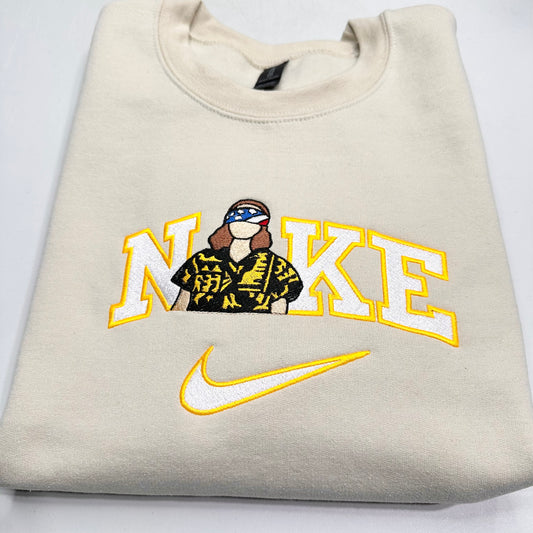 A folded cream-colored sweatshirt featuring a custom graphic of the word 'NIkE' in yellow and white varsity letters, with a stranger things eleven figure wearing a black and yellow patterned shirt and a flag headband. Below the text is a yellow and white embroidered Nike swoosh.