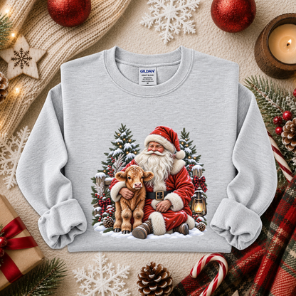 A light gray crewneck sweatshirt featuring a festive Christmas graphic of Santa Claus hugging a brown calf, surrounded by pine trees, a lantern, and snow. The sweatshirt is styled in a cozy holiday flat-lay arrangement with red ornaments, a candle, pinecones, candy canes, and a plaid scarf.