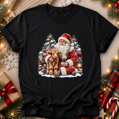 A black short-sleeve crewneck t-shirt featuring a festive graphic of Santa Claus hugging a small brown calf, surrounded by snow-covered pine trees and lanterns. The shirt is displayed in a styled flat-lay setting with Christmas decorations, including pine branches, a wrapped gift, and a red plaid scarf.