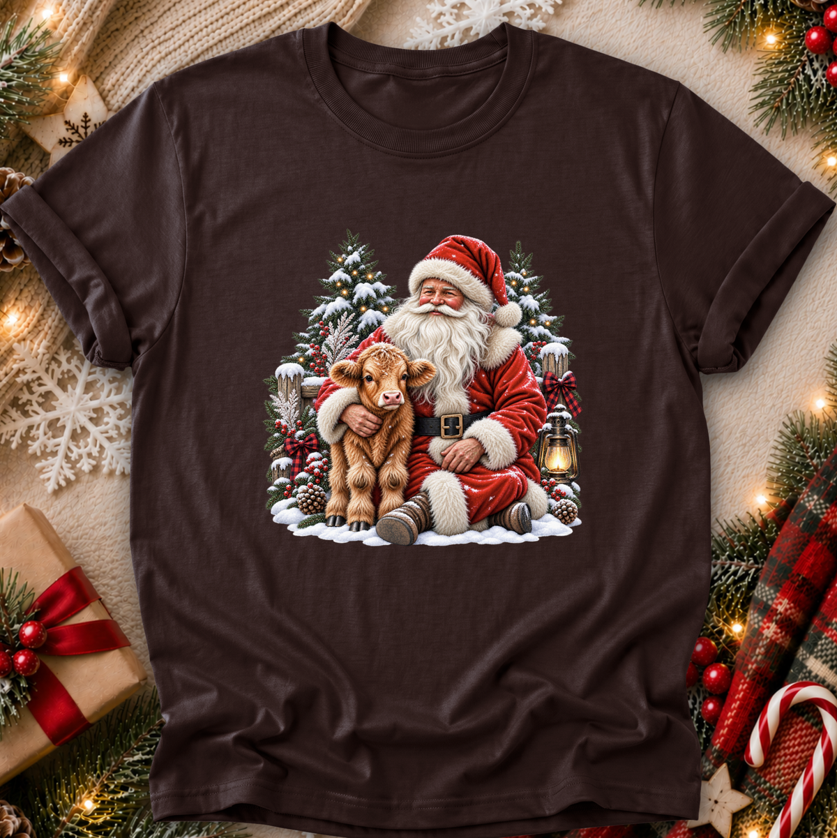 A dark brown short-sleeve t-shirt with a festive Christmas graphic of Santa Claus hugging a small brown calf, surrounded by snow, pine trees, and a lantern. The shirt is displayed in a flat-lay setting with holiday decorations like pine branches, a wrapped gift, and a plaid scarf.