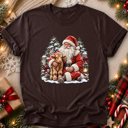 A dark brown short-sleeve t-shirt with a festive Christmas graphic of Santa Claus hugging a small brown calf, surrounded by snow, pine trees, and a lantern. The shirt is displayed in a flat-lay setting with holiday decorations like pine branches, a wrapped gift, and a plaid scarf.