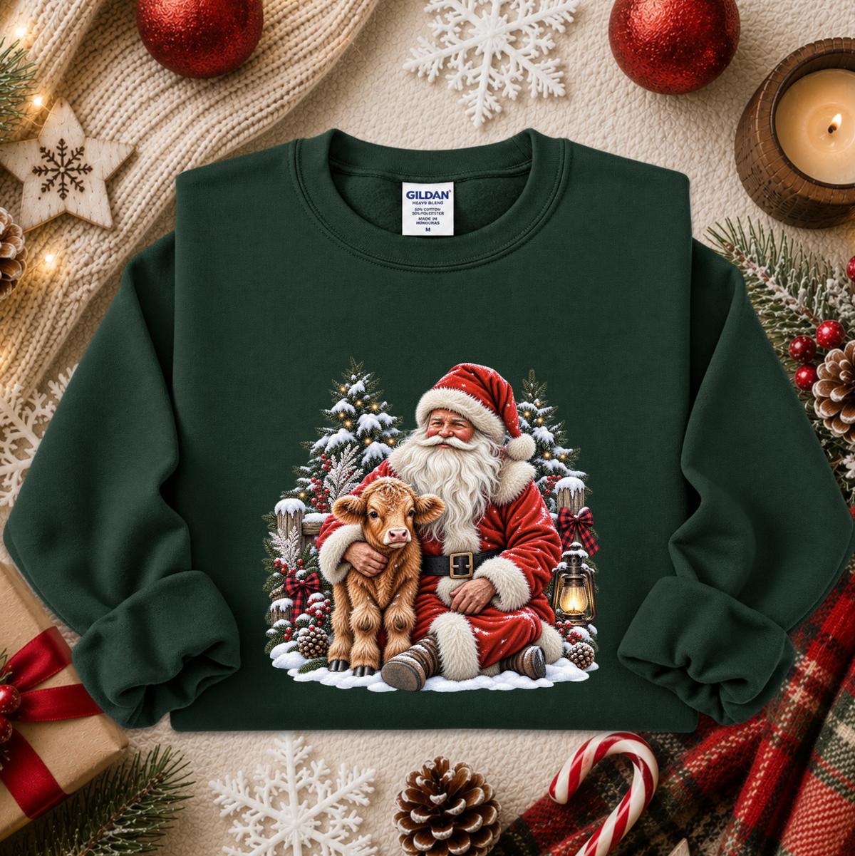 A dark forest green crewneck sweatshirt featuring a festive Christmas graphic of Santa Claus hugging a brown calf in a snowy tree scene. The sweatshirt is styled in a cozy holiday flat-lay with a knitted scarf, red ornaments, a candle, pinecones, and candy canes.