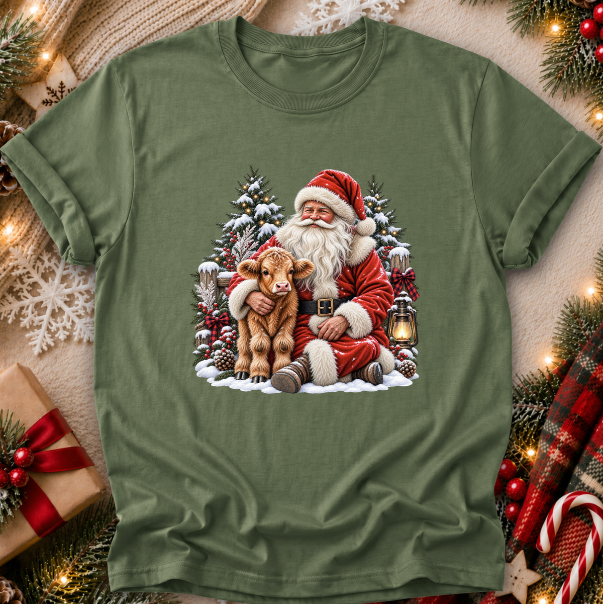 An olive green short-sleeve t-shirt with a festive Christmas graphic of Santa Claus hugging a brown calf, surrounded by snow, pine trees, and a lantern. The shirt is displayed in a flat-lay arrangement with holiday decorations like a wrapped gift, pine branches, and lights.