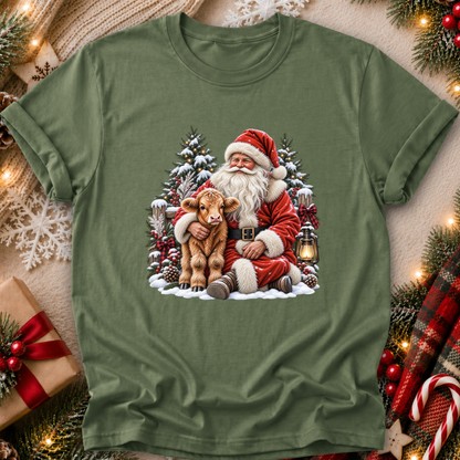 An olive green short-sleeve t-shirt with a festive Christmas graphic of Santa Claus hugging a brown calf, surrounded by snow, pine trees, and a lantern. The shirt is displayed in a flat-lay arrangement with holiday decorations like a wrapped gift, pine branches, and lights.