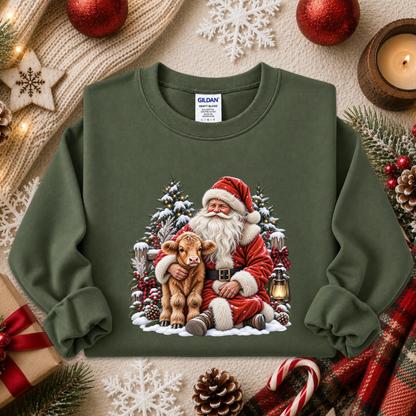 An olive green crewneck sweatshirt featuring a festive Christmas graphic of Santa Claus hugging a brown calf in a snowy forest. The sweatshirt is styled in a cozy holiday flat-lay arrangement with red ornaments, a candle, pinecones, and a gift box.