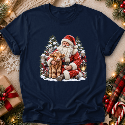 A navy blue short-sleeve crewneck t-shirt featuring a festive Christmas graphic of Santa Claus hugging a small brown calf in a snowy winter scene. The t-shirt is displayed in a flat-lay setting surrounded by holiday decorations including pine branches, a wrapped gift box, a red plaid scarf, and warm string lights.