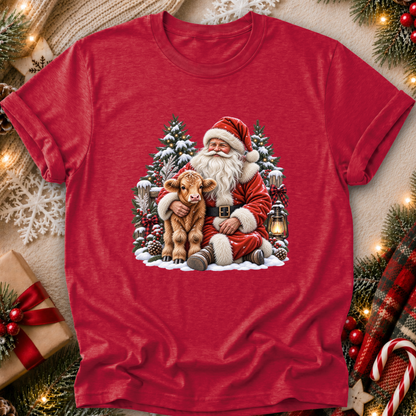 A red short-sleeve crewneck t-shirt featuring a festive Christmas graphic of Santa Claus hugging a small brown calf in front of snow-covered trees. The shirt is displayed in a styled flat-lay setting with holiday decorations including pine branches, a wrapped gift, and a plaid scarf.