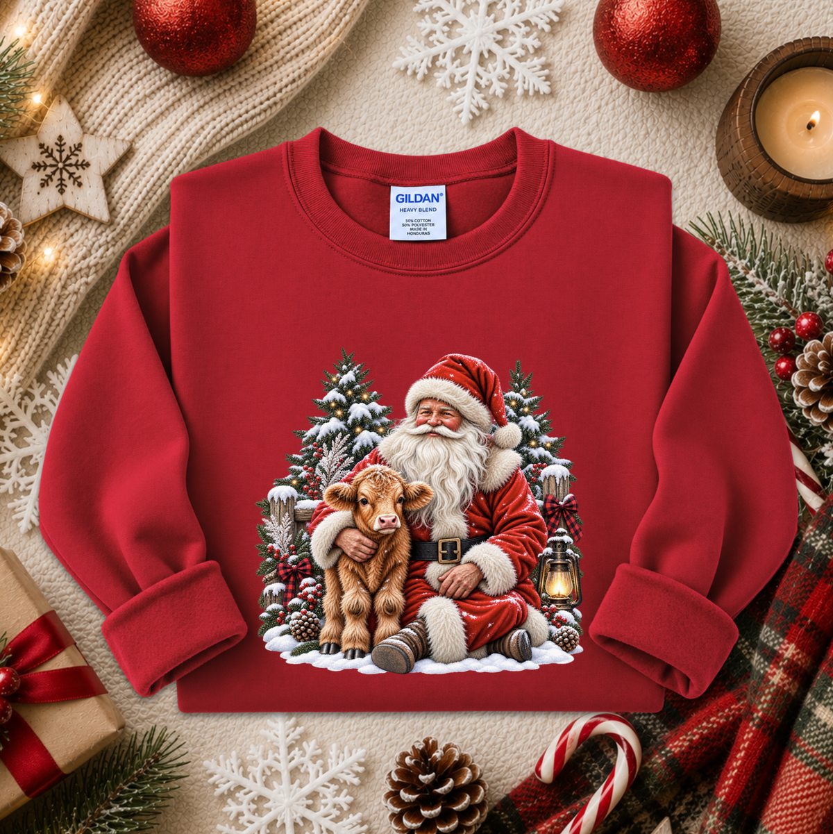 A red crewneck sweatshirt featuring a festive Christmas graphic of Santa Claus hugging a calf, surrounded by snow-covered pine trees and a lantern. The sweatshirt is displayed in a styled flat-lay setting with holiday decorations including a candle, pinecones, candy canes, and a gift box.