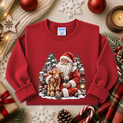 A red crewneck sweatshirt featuring a festive Christmas graphic of Santa Claus hugging a calf, surrounded by snow-covered pine trees and a lantern. The sweatshirt is displayed in a styled flat-lay setting with holiday decorations including a candle, pinecones, candy canes, and a gift box.