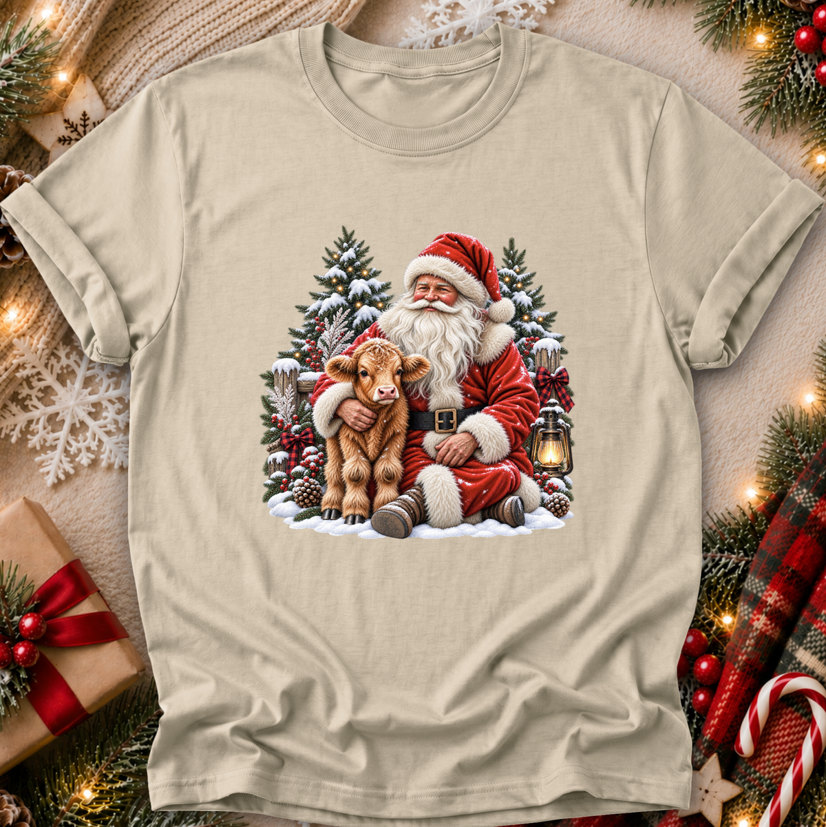 A beige crewneck t-shirt featuring a festive Christmas graphic of Santa Claus hugging a brown calf, surrounded by snow-covered pine trees and a lantern. The shirt is displayed in a cozy holiday flat-lay setting with a gift box, knitted sweater, and pine garlands.