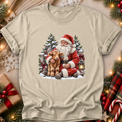 A beige crewneck t-shirt featuring a festive Christmas graphic of Santa Claus hugging a brown calf, surrounded by snow-covered pine trees and a lantern. The shirt is displayed in a cozy holiday flat-lay setting with a gift box, knitted sweater, and pine garlands.