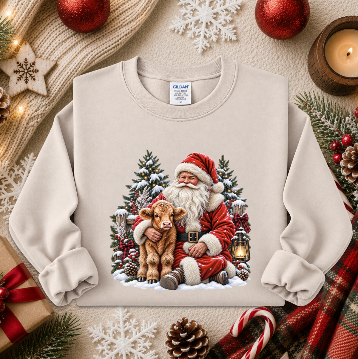 A cream-colored crewneck sweatshirt featuring a festive Christmas graphic of Santa Claus hugging a small brown calf, surrounded by snow, evergreen trees, and lanterns. The sweatshirt is styled in a cozy holiday flat-lay arrangement with a knitted sweater, pinecones, candy canes, and red ornaments.