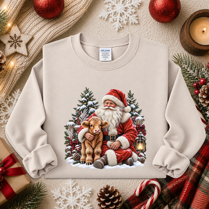 A cream-colored crewneck sweatshirt featuring a festive Christmas graphic of Santa Claus hugging a small brown calf, surrounded by snow, evergreen trees, and lanterns. The sweatshirt is styled in a cozy holiday flat-lay arrangement with a knitted sweater, pinecones, candy canes, and red ornaments.