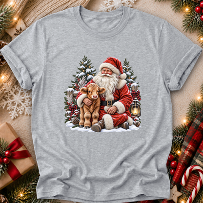 A heather gray short-sleeve t-shirt with a festive Christmas graphic of Santa Claus hugging a brown calf in a snowy winter scene. The t-shirt is displayed in a cozy flat-lay setting surrounded by holiday decorations including pine branches, a wrapped gift box, a red plaid scarf, and warm string lights.