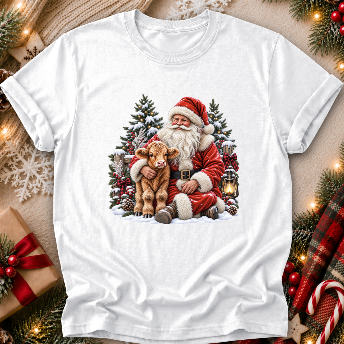 A white short-sleeve crewneck t-shirt featuring a festive Christmas graphic of Santa Claus hugging a brown calf, surrounded by pine trees, a lantern, and snow. The shirt is displayed in a styled flat-lay setting with holiday decorations like a wrapped gift, pine branches, and warm string lights.