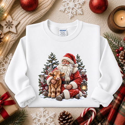 A white long-sleeve crewneck sweatshirt with a colorful Christmas graphic of Santa Claus hugging a calf, surrounded by pine trees and snow. The sweatshirt is displayed in a flat-lay arrangement with holiday decorations including red ornaments, a candle, pinecones, and candy canes.