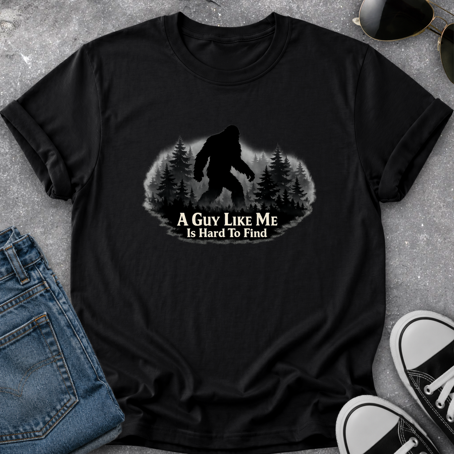 A black short-sleeve graphic t-shirt laid flat on a gray textured surface, styled with blue jeans, sunglasses, and black-and-white sneakers. The shirt features a graphic of a Bigfoot silhouette in a pine forest with the text 'A GUY LIKE ME Is Hard To Find'.